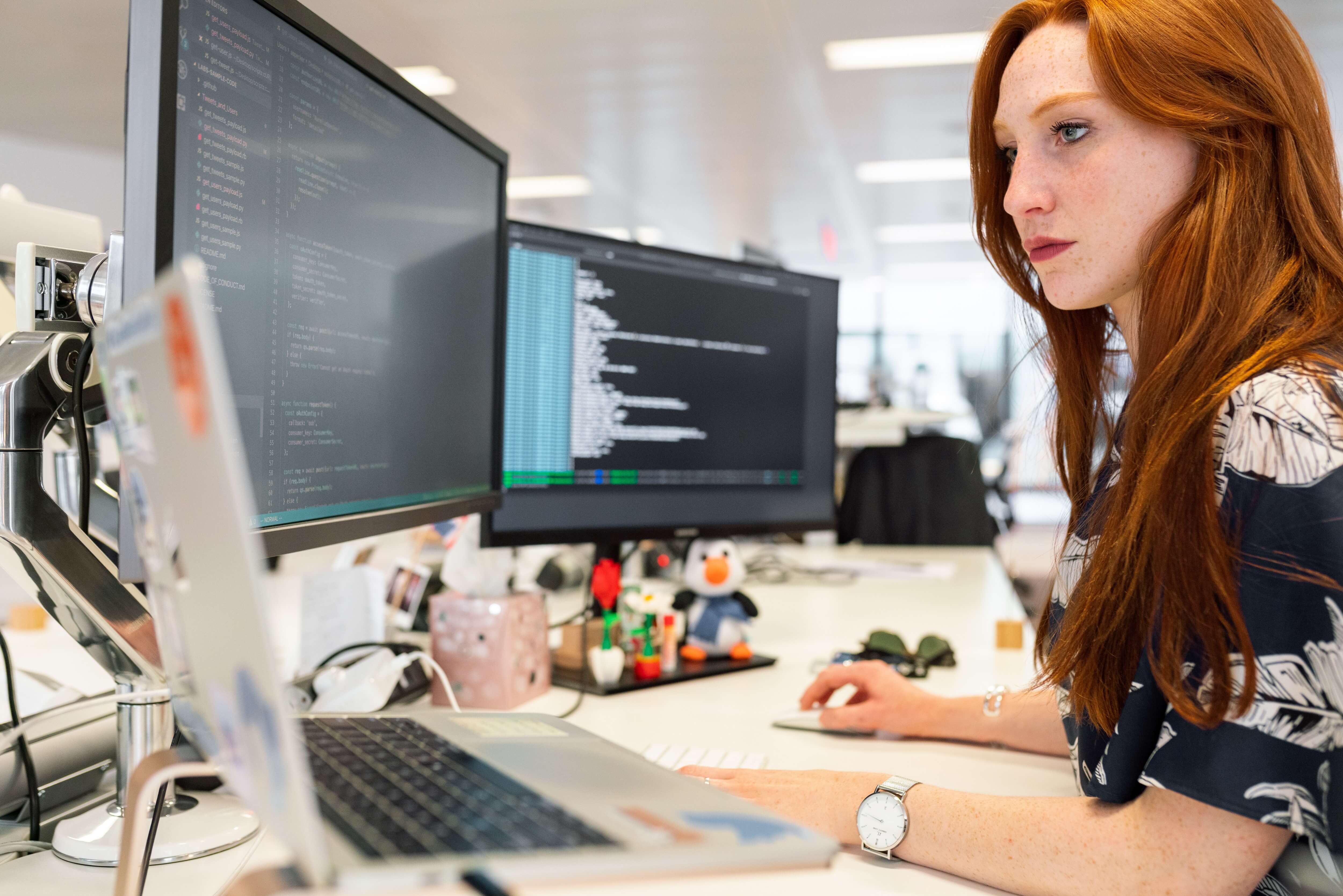 Female developer in blue floral shirt working on code with three monitors